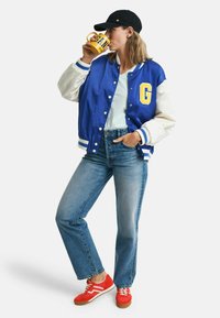 Blå og hvid varsity jakke med et gult G, lyseblå t-shirt, denim jeans og røde sneakers, holder en gul krus.