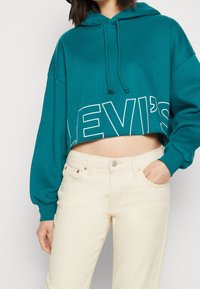 Hoodie courte couleur sarcelle avec cordon de serrage, affichant un grand logo blanc "LEVI'S". Associé à un jean droit de couleur claire. Texture en coton lisse.