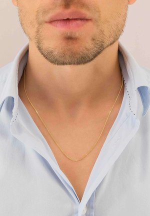 Collana di oro che riposa su pelle nuda, caratterizzata da un design sottile e liscio. Indossata sopra una camicia azzurro chiaro non abbottonata con una texture morbida.