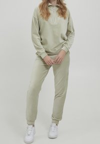 Tuta in velluto verde chiaro caratterizzato da un colletto alto con zip, polsini a coste e pantaloni affusolati coordinati, abbinati a sneakers bianche.