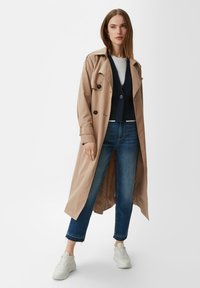 Beiger Trenchcoat mit tailliertem Gürtel, über einem marineblauen Cardigan getragen, kombiniert mit blauen Cropped-Jeans und weißen Sneakers. Glatte Stofftextur.