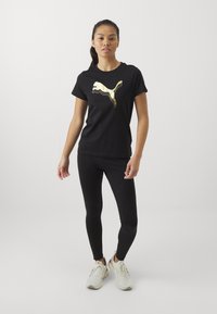 Tricou din bumbac negru cu un logo Puma auriu, asortat cu colanți negri și adidași albi. Design simplu, cu mâneci scurte și formă ajustată.