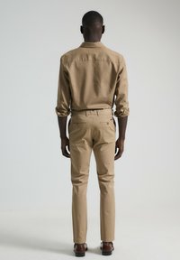 Camisa beige de botones con mangas remangadas, combinada con pantalones beige de corte slim. Zapatos de cuero marrón completan el conjunto. Diseño minimalista.