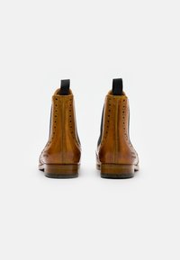 Botas Chelsea de cuero marrón con paneles elásticos laterales, detalles de acentuación tipo brogue y lengüetas. Textura suave y tacones de madera bajos.