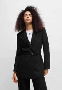Černý blazer s texturovanou látkou, dlouhými rukávy a páskem v pase. Nosený přes bílou košili s jemným logem na výstřihu.