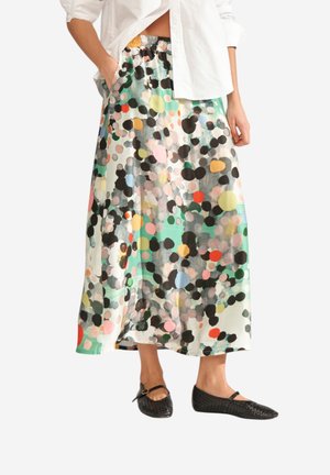 Naine kannab mitmevärvilist täpilist midi-seelikut, valget särki ja musti kootud Mary Jane tüüpi kingi, seisab käed taskutes.