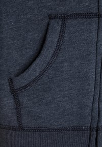 Donkerblauwe sweatshirtstof met een zachte textuur, met een ronde zak en contrasterende stiksels langs de randen.