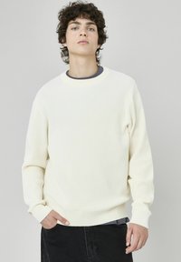 Pull à côtes couleur crème avec un col ras du cou et des manches longues, présentant une surface texturée et un ourlet ajusté, porté par-dessus une couche de base sombre.