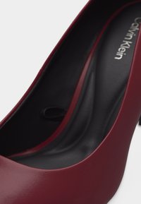 Γυναικείο παπούτσι με ψηλό τακούνι από δέρμα σε μπορντό χρώμα, με μαύρη επενδυμένη εσωτερική σόλα και λευκό λογότυπο Calvin Klein τυπωμένο στην περιοχή της φτέρνας.
