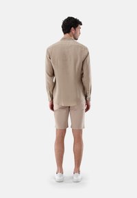 Helles beigefarbenes Leinenhemd mit Kragen und langen Ärmeln, mit Knopfmanschetten, kombiniert mit beigen Shorts und weißen Turnschuhen. Rückansicht gezeigt.