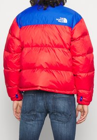 Veste matelassée isolante rouge et bleue avec une finition brillante, arborant un logo dans le haut du dos et des accents bleus aux poignets.