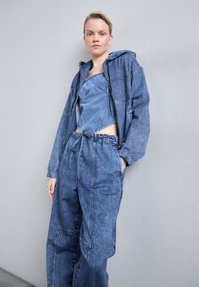 Denim apģērbs, kas sastāv no apgrieza jaku ar rāvējslēdzēju un platu biksēm, abi ir dažādās zilo toņos ar redzamu šuvumu un brīvu piegriezumu.