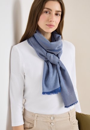 Mladá žena s dlouhými hnědými vlasy nosí bílý top a modrý šátek uvázaný volným uzlem, stojí na světlém pozadí.