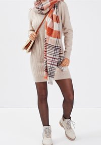 Strickkleid in Beige mit strukturiertem Muster, kombiniert mit einem orangefarbenen karierten Schal und beige Schnürstiefeln mit braunen Akzenten.