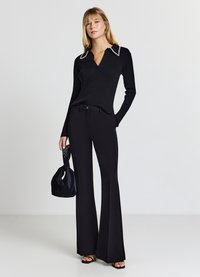 Top nero a maglia a coste con scollo a V e colletto con perline bianche, abbinato a pantaloni neri a zampa. Una piccola borsa nera è tenuta in una mano.