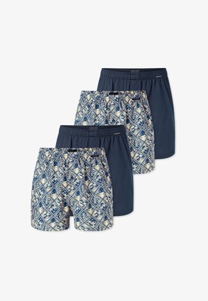 Hombre con los brazos cruzados, vistiendo bañador estampado en azul y beige, de pie frente a un fondo blanco liso.