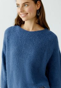 Pull ample, col rond en tissu bleu duveteux. Présente des manches longues et une texture douce. Aucune motif ou accent visible.