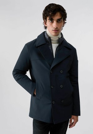 Jeune homme portant un manteau sombre croisé sur un pull à col roulé blanc, debout devant un fond clair uni.