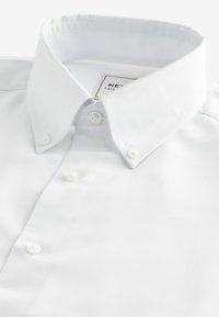 Chemise en coton blanche avec un col boutonné, dotée de deux boutons sur le devant et d'une texture lisse. Étiquette visible sous le col.