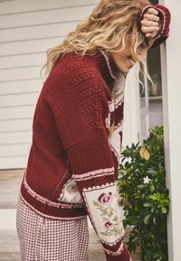Karmesinroter Strickpullover mit hohem Kragen und floralen Mustern auf den Ärmeln. Getragen über karierten Hosen, mit einer strukturierten Oberfläche.