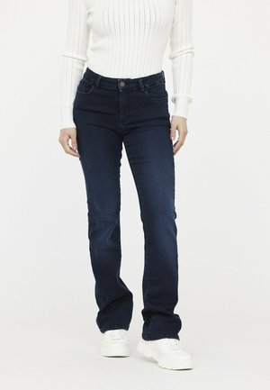 Jeans en denim bleu foncé avec une coupe droite, un design à cinq poches et une fermeture à bouton. Portés avec un pull en maille côtelée blanche et des baskets blanches.