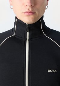 Sort lynlåsjakke med høj krave, med hvide syninger og et lille "BOSS" logo på venstre brystområde.