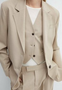 Completo beige a tre pezzi composto da giacca, gilet e pantaloni, caratterizzato da una superficie liscia e bottoni scuri. Design di sartoria classica.