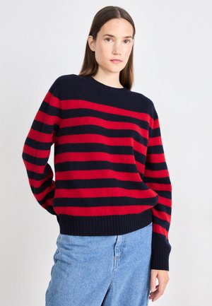 Czerwono-granatowy sweter w paski z okrągłym dekoltem, ściągaczami przy rękawach i u dołu, wykonany z miękkiej tkaniny, noszony do jasnoniebieskich dżinsów.