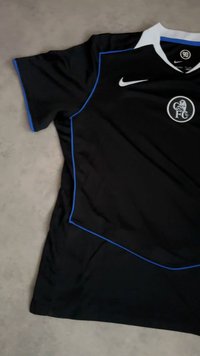 Black Chelsea FC soccer jersey with white collar, blue trim, Nike logo, and club crest on chest, laid flat on gray surface.