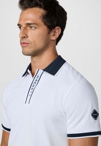 Biela polo košeľa s námorníckym golierom a námorníckymi akcentmi. Obsahuje logo na ľavom rukáve a podrobný zapínací panel. Hladká textúra.