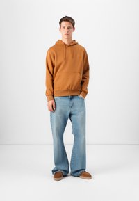Un sweat à capuche marron avec une poche avant, associé à un jean large bleu clair et des chaussures slip-on marron. Design simple et décontracté.