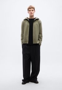 Φούτερ με φερμουάρ σε ελιά πράσινο χρώμα και λογότυπο στο μπροστινό μέρος, φορεμένο πάνω από μαύρο πουκάμισο. Συνδυάζεται με άνετο μαύρο παντελόνι και μαύρα παπούτσια. Απλός, μοντέρνος σχεδιασμός.