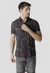 Hombre vistiendo una camisa de manga corta negra con botones y estampados coloridos de flores y animales, ajustándose el cuello, de pie frente a un fondo gris.