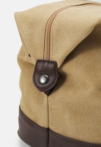 Tan canvas bag with a brown leather base, featuring a folded top, zipper closure, and a snap button accent. Textured surface and smooth hardware.