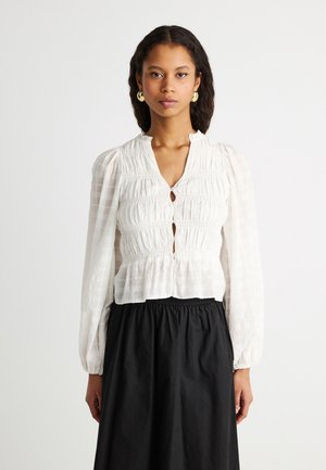 Lokkis juustega naine kannab valget tekstuuriga pika varrukaga pluusi eespool nuppudega ja musta kõrge vöökohaga seelikut ühetoonilise tausta ees.