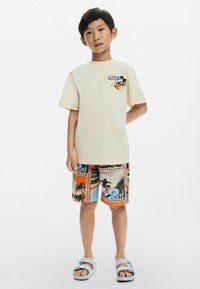 Svijetlo žuta majica s crtanom grafikom na prsima, u kombinaciji s višebojnim šortsevima koji imaju uzorke na temu surfanja i plaže. Plave sandale.