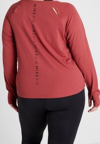Lange mouw sporttop in baksteenrood, met een gestructureerd patroon, uitsnede aan de achterkant en een zwart "Nike Air" logo verticaal aan de zijkant.