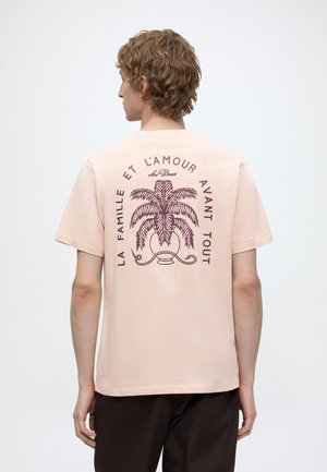 Homme aux cheveux bouclés vu de dos portant un t-shirt beige avec un palmier et un texte en français au dos.