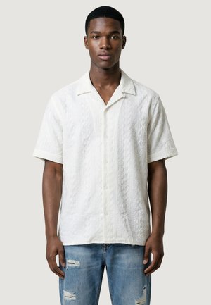 Młody mężczyzna ubrany w białą, krótki rękaw, teksturowaną koszulę zapinaną na guziki oraz podniszczone jasnoniebieskie jeansy, stoi na tle gładkiego tła.