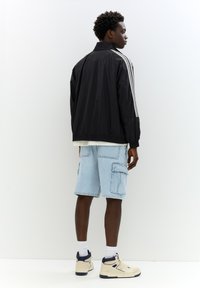 Sort letvægtsjakke med hvide striber på ærmerne, parret med lyseblå cargo shorts og beige høj-top sneakers.