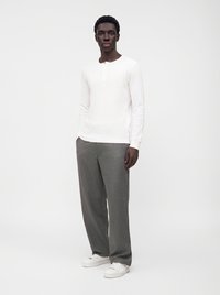 Homme debout portant une chemise blanche à manches longues, un pantalon gris ample et des baskets blanches sur un fond clair uni.