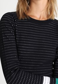 Pull noir côtelé orné de pois blancs, col rond, manches longues et poignets contrastants blancs et bleu sarcelle.