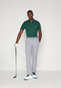 Πόλο μπλούζα σε σκούρο πράσινο, ανοιχτό γκρι παντελόνι, λευκά παπούτσια γκολφ με μπλε λεπτομέρειες, κρατώντας ένα μπαστούνι γκολφ, μινιμαλιστικό φόντο.