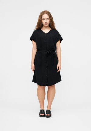 Kobieta z długimi rudymi włosami w czarnej sukience na guziki z paskiem w talii i czarnych, otwartych sandałach wsuwanych, na jednolitym tle.