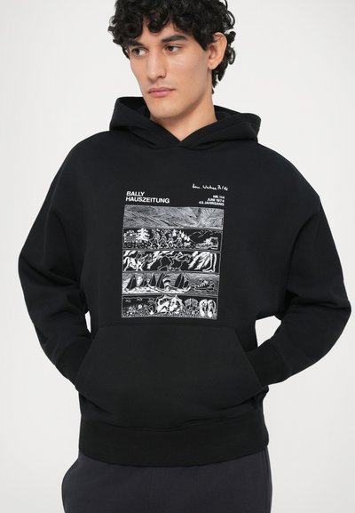 Giovane uomo che indossa un hoodie nero con pannelli grafici bianchi che raffigurano scene della natura e la scritta "Bally Hauszeitung."