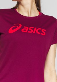 Camiseta atlética magenta fabricada con tejido elástico, que presenta un cuello redondo y un gran logo rojo de ASICS en el pecho.
