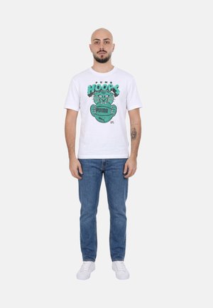 Uomo che indossa una t-shirt grafica bianca Puma Hoops, jeans blu e scarpe da ginnastica bianche, in piedi rivolto in avanti con espressione neutra.
