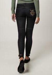 Tmavě modré džínsy s úzkým střihem, s dekorativní zlatou výšivkou na zadní kapse a zipovými detaily na kotnících.