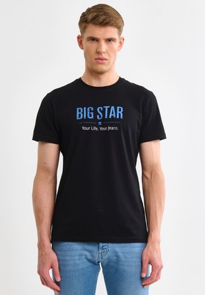Joven vestido con una camiseta negra que dice "BIG STAR Your Life, Your Jeans" y jeans azules, de pie frente a un fondo blanco liso.