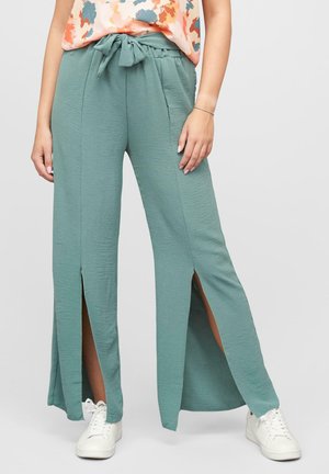 Lichtgroene, wijde pantalon met een strik taille, voorzien van verticale inkepingen aan de zijkanten en een textuurstof. G gedragen met witte sneakers.